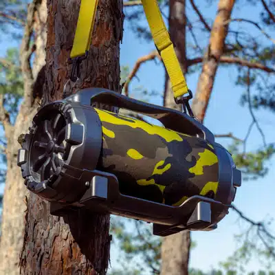 Large portable Bluetooth speaker with camouflage cover and yellow strap hanging from a tree outdoors
