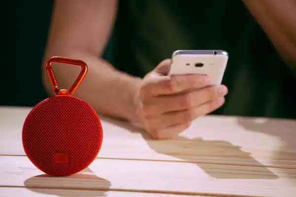 A small red portable Bluetooth speaker sits on a wooden table beside someone holding a smartphone