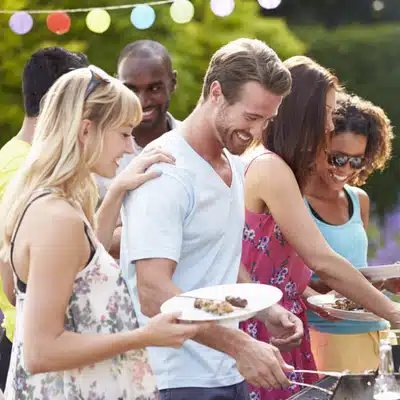 A group of friends smiling and cooking food together at an outdoor barbecue gathering in daylight