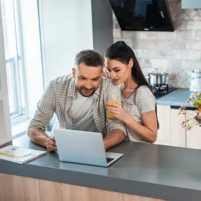 Couple leans over laptop in kitchen, researching what is a smarthome and planning upgrades together