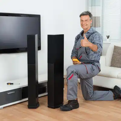 Installer kneels beside tower speakers near television, giving thumbs-up after successful home audio setup completed