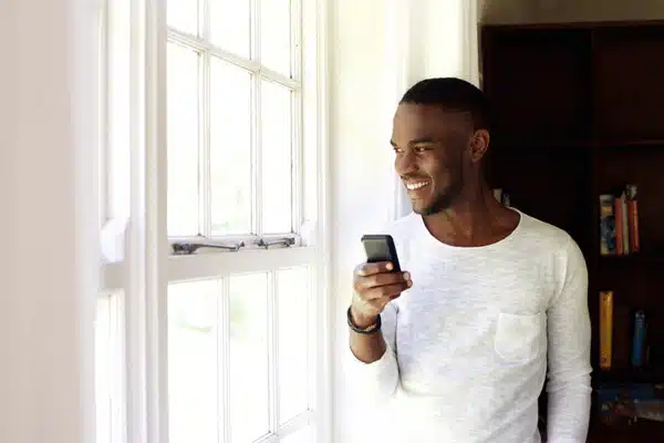 Smiling man stands by bright window, checking smartphone notifications at home, relaxed and content today