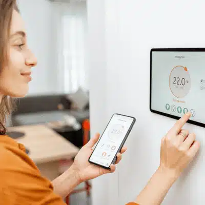 Woman adjusts wall tablet thermostat while holding phone, managing home temperature and energy efficiently remotely