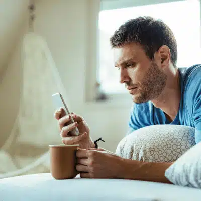 Relaxed man holds smartphone and mug on couch, reading messages and checking home notifications quietly