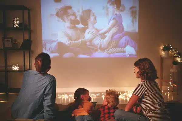 Family watches movie projected on wall with string lights, enjoying cozy home theater evening together