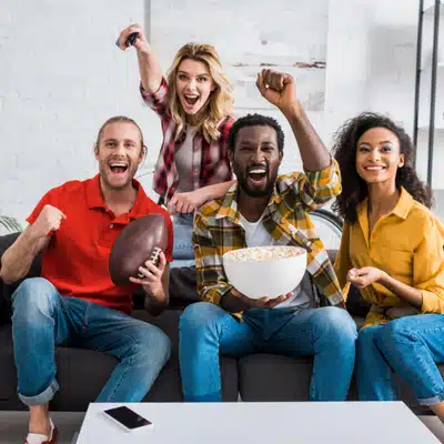 Friends cheer on couch watching sports together, snacks ready, celebrating exciting play at home loudly