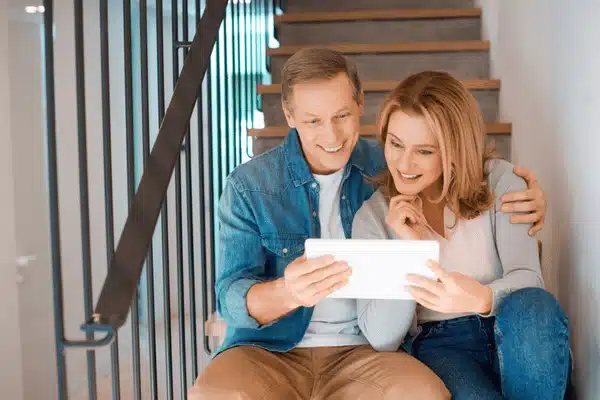 Smiling couple sits on stairs, sharing tablet while browsing photos inside bright modern home together