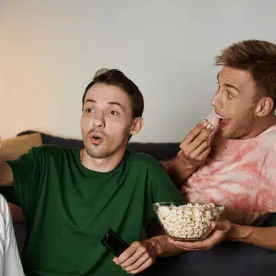Two friends watch television, reacting with excitement while sharing popcorn on a cozy couch together