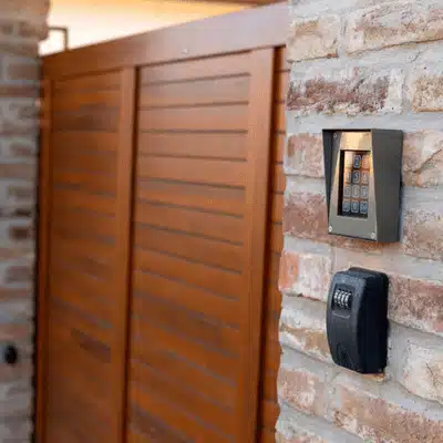 Outdoor keypad and reader mounted on brick by wooden gate; residential intercom system controls access