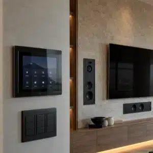 A modern living room features a wall-mounted touchscreen, lighting keypads, speakers, and television for a control4 vs crestron automation system